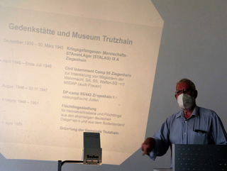 Mann mit Maske und Brille, vor einer Projektionswand mit Text zur Gedenkstätte Trutzhain und historischen Ereignissen.