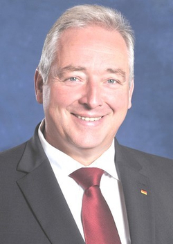 Lächelnder Mann im Anzug mit roter Krawatte und deutscher Flaggennadel.