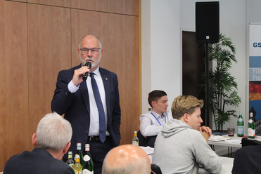 Ein Mann im Anzug mit Brille spricht in ein Mikrofon bei einer Konferenz, mit Zuhörern im Vordergrund und einem Banner im Hintergrund.