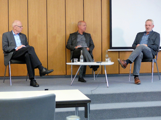 Drei Männer sitzen auf einer Bühne bei einer Podiumsdiskussion, mit einem Tisch, Wasserflaschen und einem großen Bildschirm im Hintergrund.