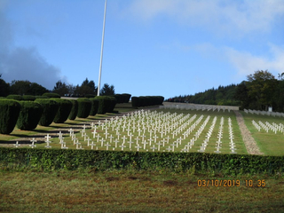 Militärfriedhof mit unzähligen weißen Kreuzen auf grünem Hügel. Hecken, Bäume und Fahnenmast unter blauem Himmel.