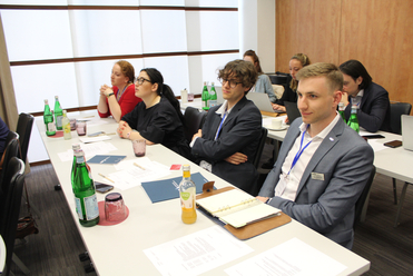 Personen, darunter Männer und Frauen, sitzen an Tischen in einem hellen Konferenzraum und hören während eines Treffens zu, mit Getränken und Papieren.