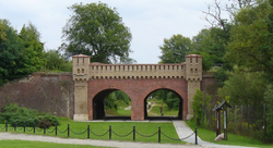Historische Ziegelbrücke mit zwei Torbögen und Türmen in einem Park. Grüne Bäume und Gras, Menschen gehen hindurch.