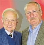 Zwei lächelnde ältere Männer, einer mit weißen Haaren in einem blau karierten Sakko, der andere mit Brille und Schnurrbart.