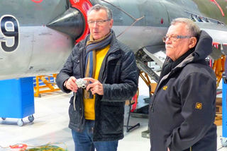 Zwei Männer, einer mit Kamera, stehen in einem Hangar oder Museum neben einem grauen Düsenflugzeug mit der Nummer '9'.