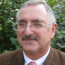 Porträt eines älteren Mannes mit Brille und Schnurrbart, brauner Jacke, leicht lächelnd, vor grünem Hintergrund.