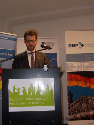 Mann spricht an einem Rednerpult vor Bannern der Konrad-Adenauer-Stiftung, GSP und Haus der Brandenburgisch-Preußischen Geschichte.
