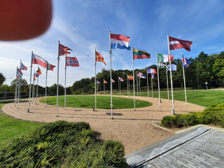 Vielfältige Nationalflaggen an Masten in kreisförmiger Anordnung auf Schotter, umgeben von Rasen und Bäumen, blauer Himmel.