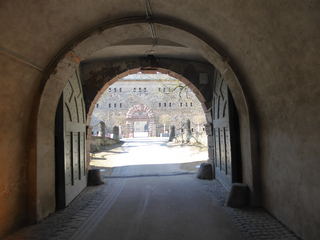 Blick aus einem dunklen, gewölbten Steintunnel mit offenen Holztoren auf eine sonnenbeschienene Festungsmauer mit einem verzierten Torbogen.