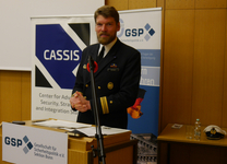 Ein bärtiger Mann in Marineuniform steht hinter einem Rednerpult vor Bannern mit GSP und CASSIS Logos.