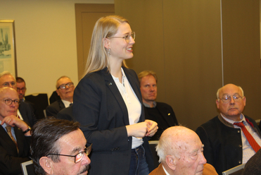 Lächelnde blonde Frau mit Brille spricht stehend, umgeben von sitzenden älteren Männern in einem Besprechungsraum.