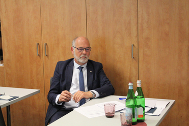 Ein bärtiger Mann mit Brille in Anzug sitzt an einem Tisch mit Wasserflaschen, Gläsern und Papieren vor einer Holzwand.