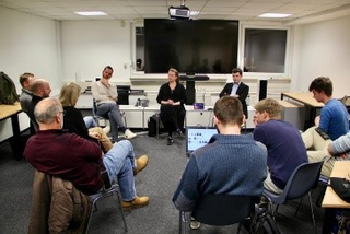 Menschen bei Diskussion/Seminar; Frau und Mann sprechen im Zentrum, großer Bildschirm dahinter.