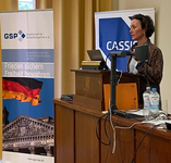 Frau spricht an Rednerpult, hinter ihr Banner der GSP ('Frieden sichern Freiheit bewahren') mit Reichstag und deutscher Flagge.