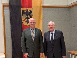 Zwei lächelnde ältere Männer in Anzügen stehen vor einer deutschen Flagge mit dem Bundesadler.