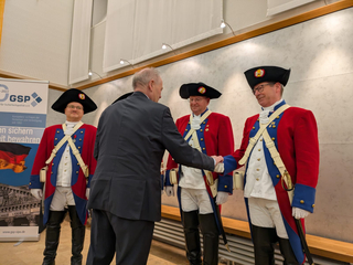 Ein Mann im Anzug schüttelt die Hand eines Mannes in historischer Uniform (roter Mantel, Dreispitz); zwei weitere stehen daneben.