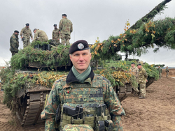 Ein lächelnder deutscher Soldat in Uniform steht vor einem mit Ästen getarnten Kampfpanzer mit weiteren Soldaten im Hintergrund.