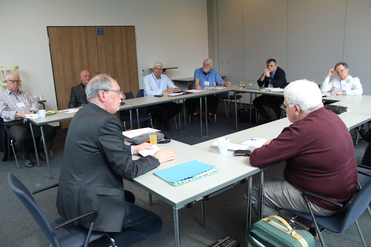 Gruppe älterer Männer bei einer Konferenz oder einem Meeting an einem U-förmigen Tisch in einem hellen Raum.