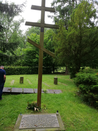 Orthodoxes Kreuz auf einer grünen Wiese, umgeben von Bäumen. Im Vordergrund Gedenktafeln für sowjetische Kriegsgefangene.