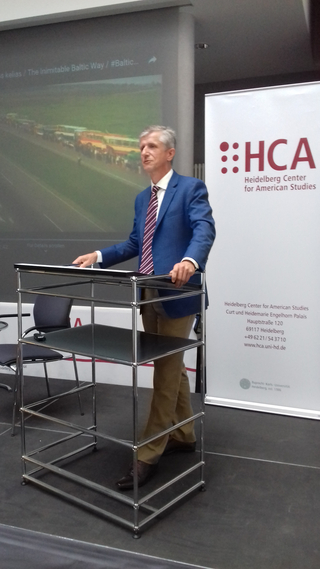 Ein Mann spricht an einem Pult vor einer Leinwand mit „The Inimitable Baltic Way“ und einem Banner des HCA.