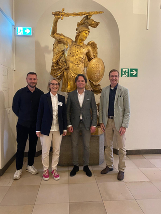 Vier Personen, darunter eine Frau im Blazer mit pinken Schuhen, posieren lächelnd vor einer goldenen Erzengel-Michael-Statue.