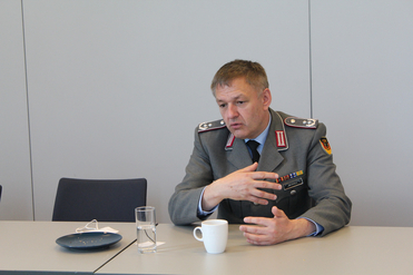 Ein deutscher Offizier in Uniform sitzt am Tisch, gestikuliert sprechend. Tasse, Wasserglas und Teller stehen bereit.
