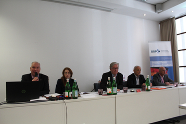 Fünf Personen, darunter einer mit Mikrofon, sitzen an einem langen weißen Tisch während einer Sicherheitspolitik-Konferenz.