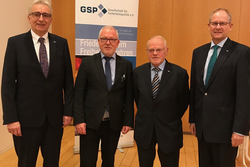 Vier Männer in Anzügen posieren vor einem Banner der GSP (Gesellschaft für Sicherheitspolitik e.V.).