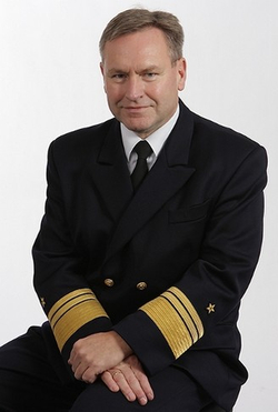Porträt eines Mannes in dunkler Marineuniform mit goldenen Rangstreifen an den Ärmeln, der in die Kamera blickt.