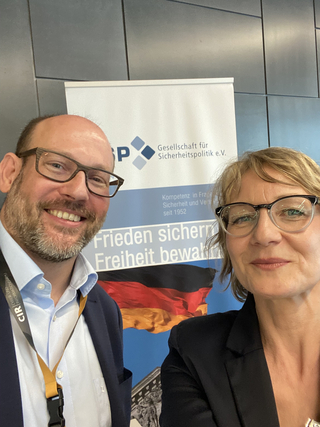 Lächelnder Mann und Frau machen ein Selfie vor einem Banner der Gesellschaft für Sicherheitspolitik mit deutscher Flagge.