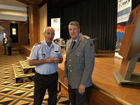 Zwei Männer in Uniformen, einer im hellblauen Hemd, einer in grauer Militäruniform, stehen auf einer Bühne mit Eventbannern.