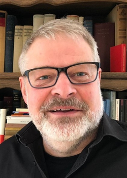 Porträt eines lächelnden Mannes mit Brille und grauem Bart vor einem Bücherregal.