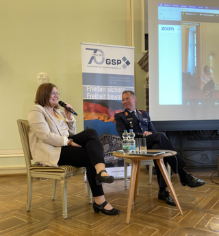Eine Frau spricht mit Mikrofon und ein Mann in Militäruniform sitzt ihr gegenüber, mit GSP-Banner und Projektionswand im Hintergrund.
