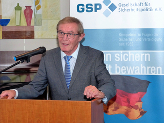 Ein Mann im Anzug spricht an einem Rednerpult vor einem Banner der Gesellschaft für Sicherheitspolitik mit deutscher Flagge.