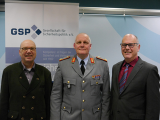 Drei Männer, darunter ein Offizier in Uniform, vor einem Banner der Gesellschaft für Sicherheitspolitik e.V.