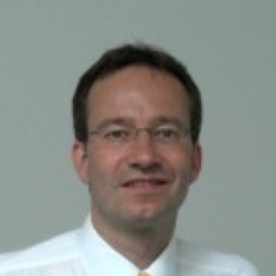 Porträt eines lächelnden Mannes mit dunklen Haaren und Brille, der ein weißes Hemd und Krawatte vor grauem Hintergrund trägt.