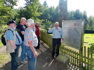 Ältere Männer bei einer Führung im Wald. Ein Führer erklärt vor einer Informationstafel die Gedenkstätte Waldfriedhof Trutzhain.