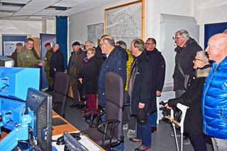 Ein Militäroffizier unterrichtet eine Gruppe Zivilisten in einem Kontrollraum, mit Karten an der Wand und Computerausrüstung.