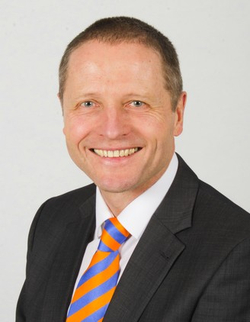 Professionelles Porträt eines lächelnden Mannes in einem dunklen Anzug, weißem Hemd und blau-orange gestreifter Krawatte.