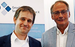 Zwei lächelnde Männer in Geschäftskleidung stehen vor einem Banner einer Sicherheits- oder Verteidigungsorganisation.