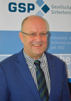 Ein lächelnder, glatzköpfiger Mann mit Brille in blauem Sakko und gestreifter Krawatte vor einem GSP-Sicherheitsbanner.
