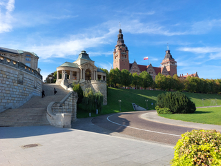 Blick auf die historischen Wały Chrobrego in Stettin mit imposanten Gebäuden, großer Treppe, grünen Hängen und polnischer Flagge unter blauem Himmel.