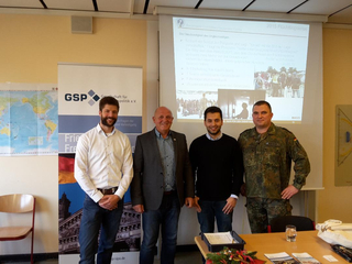 Vier Männer, darunter ein Soldat, lächeln vor einem GSP-Banner und einer Projektion zur Flüchtlingskrise 2015.