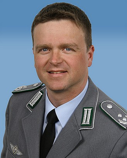 Porträt eines lächelnden Militäroffiziers in grauer Uniform mit Pilotenabzeichen, der in die Kamera blickt.