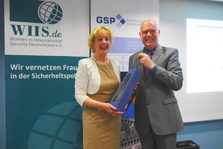 Lächelnde Frau in beigem Kleid erhält eine blaue Geschenktüte von einem Mann im Anzug bei einer Veranstaltung mit Logos.