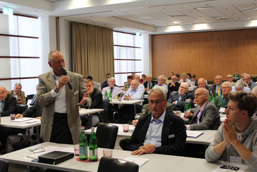 Ein Mann spricht mit einem Mikrofon vor einem Publikum von Männern, die an Tischen in einem Konferenzraum sitzen.