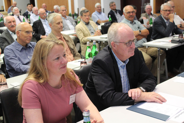 Eine Frau und mehrere Männer sitzen aufmerksam bei einer Konferenz oder einem Meeting an Tischen.