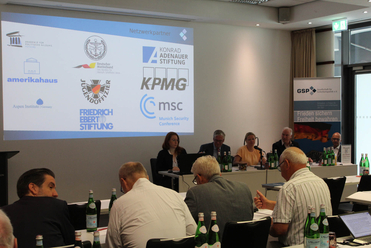 Konferenzraum mit Projektorleinwand mit Partnerlogos, einem Rednerpult und Teilnehmern an Tischen.