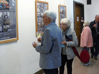 Mehrere ältere Erwachsene auf einer Innenausstellung mit gerahmten Plakaten, ein Mann hält Wein und betrachtet ein Display.