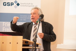 Ein älterer Mann mit grauen Haaren spricht lebhaft an einem Pult, gestikulierend mit seiner rechten Hand, GSP-Banner im Hintergrund.